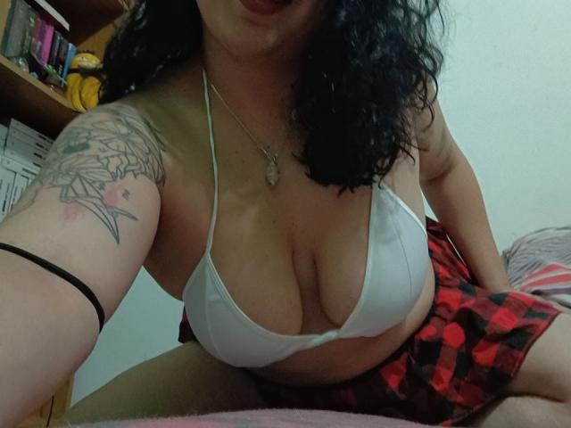 SweetYamii with curly black hair, white bikini top showing big tits, red plaid skirt, tattoo on right arm, black hair tie, and necklace, kneeling on bed.