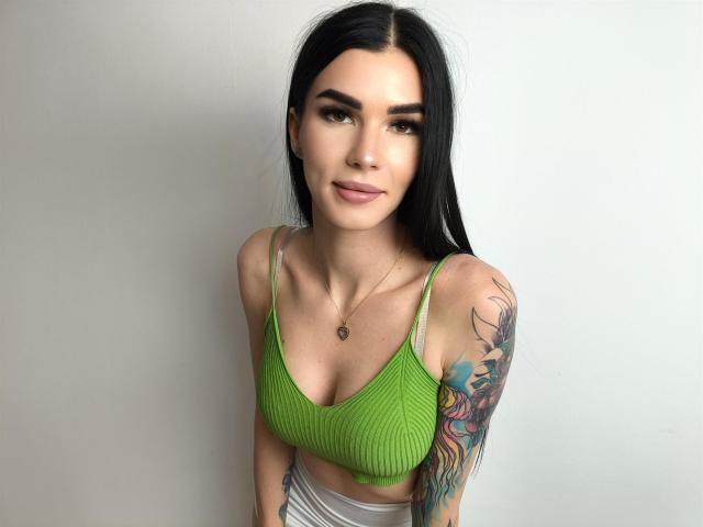 SofiaDream with long black hair, green ribbed crop top, small breasts, tattooed arm, light skin, subtle makeup, silver pants, necklace, slight smile, standing against plain white wall.
