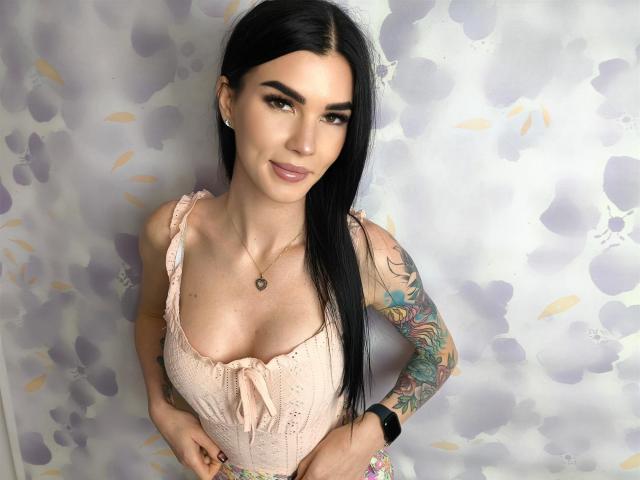 SofiaDream with black hair, wearing a pink lace top, showing her tits, tattooed arm, necklace, and wristwatch, smiling at the camera.