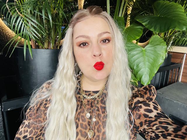 SamanthaGold with blonde wavy hair, leopard print shirt, multiple necklaces, and bright red lipstick, making a pouty face. Her ass is not visible. Her tits are average.