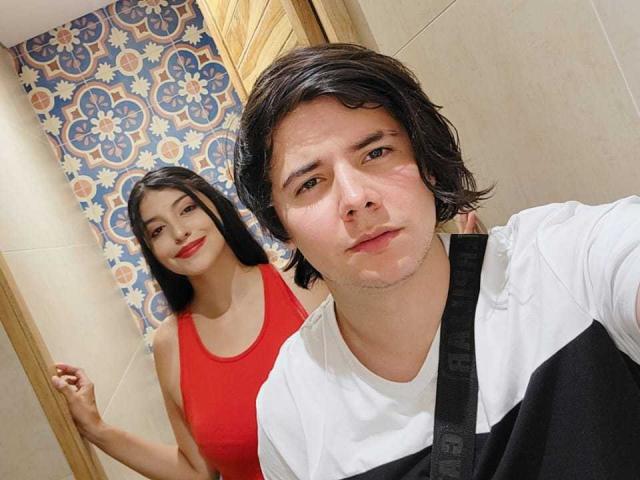 EmyAndJack with short black hair, white shirt, black straps, and Jack with medium brown hair, red top, and red lipstick, standing in a tiled room.