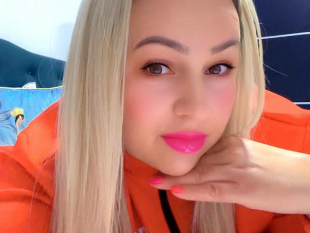 HotKrystall with blonde hair, pink lipstick, and orange hoodie, resting chin on hand, brown eyes, pink nails, soft makeup, looking at camera.