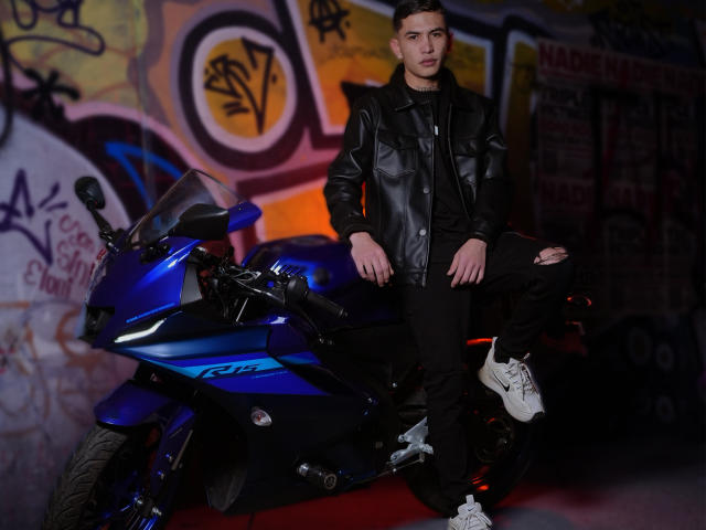 KylieAndJustin standing next to a blue Kawasaki motorcycle, wearing a black leather jacket, black ripped jeans, and white sneakers, with short brown hair and a serious expression.