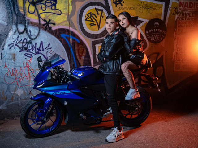 KylieAndJustin with black leather jacket, fishnet stockings, and white sneakers, sitting on a blue Yamaha motorcycle, Justin standing, both with short dark hair, in an urban setting.