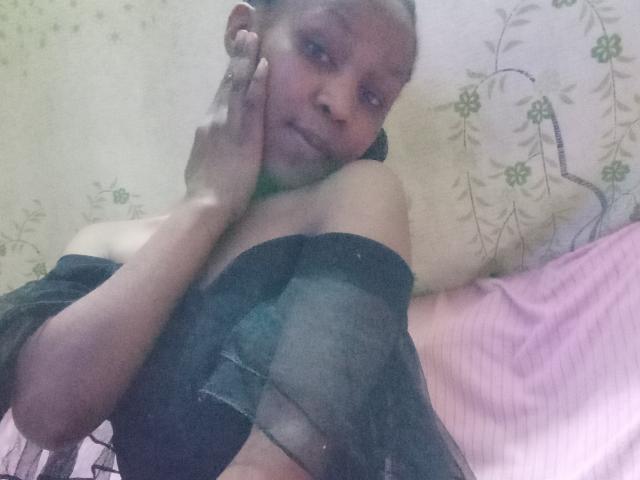 SweetCandyHotty with dark skin, small breasts, and a green choker, wearing a black sheer top, resting her hand on her cheek, looking at the camera.