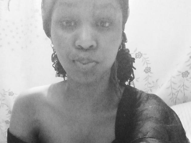 Black and white selfie of SweetCandyHotty with curly hair, dark skin, and a sheer black top, showing her bare shoulders and a subtle smile. Her tits are small.