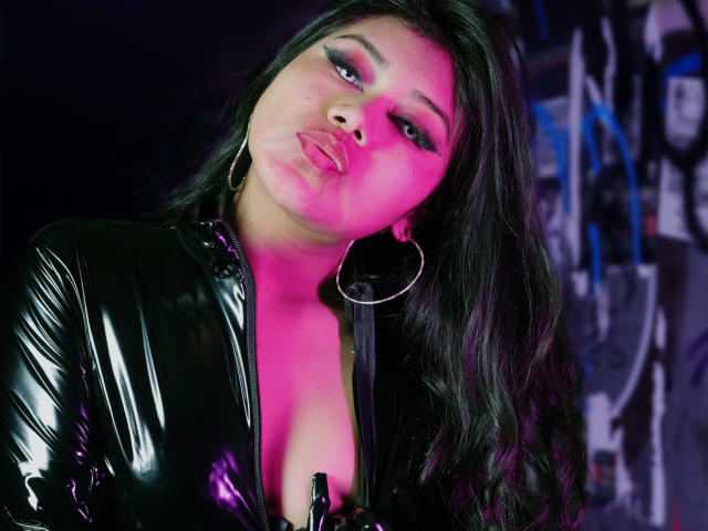 LiaDecker with long black hair, pink eyeliner, and glossy lips, wearing a shiny black latex jacket, pouting, holding a bottle, large hoop earrings, pink and purple lighting.