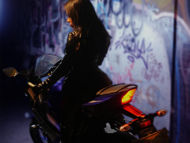 LiaDecker with long brown hair, wearing a black leather outfit, sitting on a dark purple motorcycle with a red taillight, her ass slightly raised, in a dimly lit urban setting.