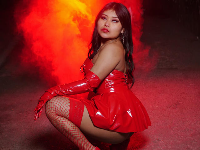 LiaDecker with red latex outfit, gloves, and fishnet stockings, squatting, red smoke behind, looking at camera, glossy skin, dark wavy hair, red lipstick, sexy, confident.