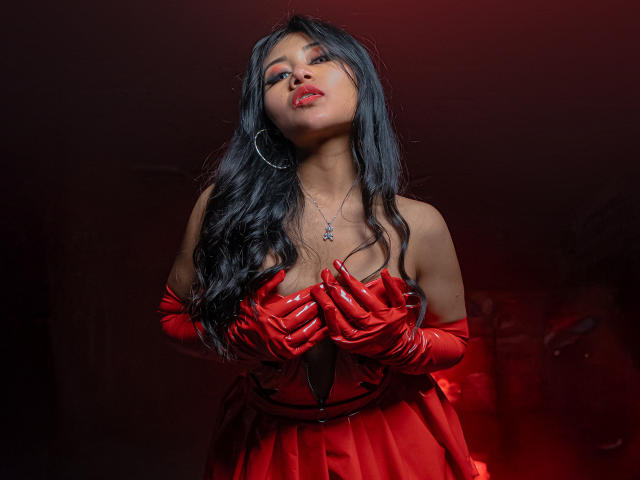LiaDecker with long black hair, red glossy latex dress, and matching gloves, holding her tits, red lipstick, hoop earring, necklace, and dark red background, looking sexy and confident.