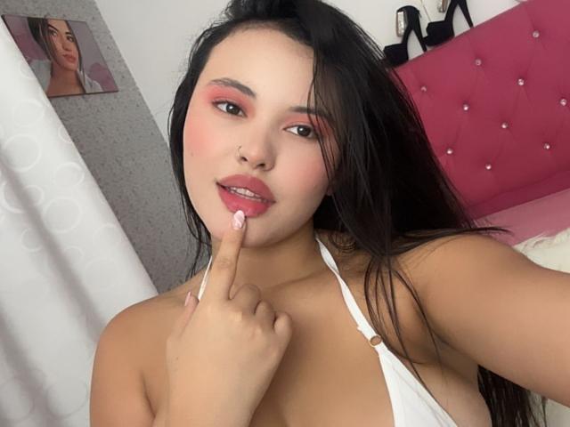 ArianaSmith69 with pink eyeshadow, nose ring, and pink lipstick, finger on her bottom lip, white halter top, showing her tits, black hair, pink bedhead in background.