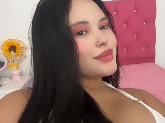 ArianaSmith69 has black hair, pink eyeshadow, a nose ring, and a white top. She's smiling with red lipstick. Her tits are small, and she's sitting on a pink bed.
