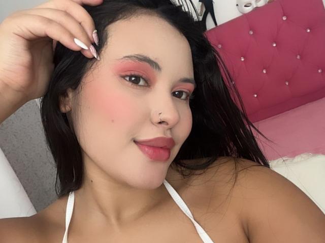 ArianaSmith69 with pink eyeshadow, white nail polish, nose ring, and dark hair, smiling at the camera, wearing a white halter top, showing her tits.