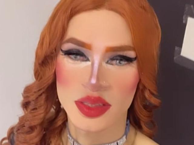 HannaTrs has red curly hair, blue eyes, pink blush, red lipstick, and thick black eyeliner. She's wearing a silver choker. Her face is slightly pixelated.