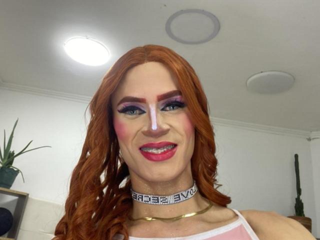 HannaTrs has red hair, pink nose, heavy makeup with purple eyeshadow, pink blush, red lipstick, and a white choker with black patterns. She's smiling with visible teeth.