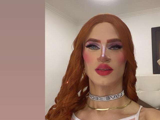 HannaTrs has red hair, heavy makeup with pink eyeshadow, red lipstick, and a white choker with black letters. She's wearing a gold necklace. Her tits are not visible.