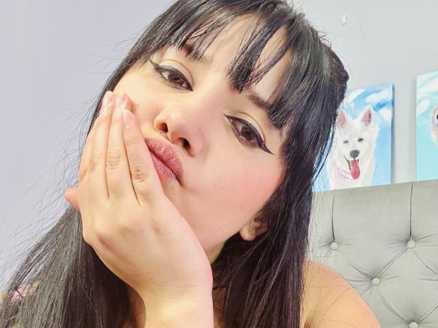 Kkatalina with black hair, pouty lips, and winged eyeliner, hand on cheek, looking at camera, gray tufted headboard, painting of white dog with pink tongue in background.