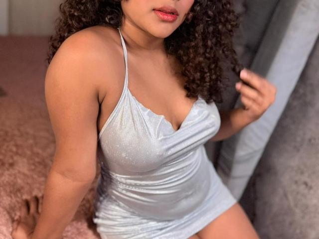 AnnyRestrepo with curly brown hair, wearing a shiny silver halter dress, showing her tits, pink lips, and tan skin, sitting on a pink bed.