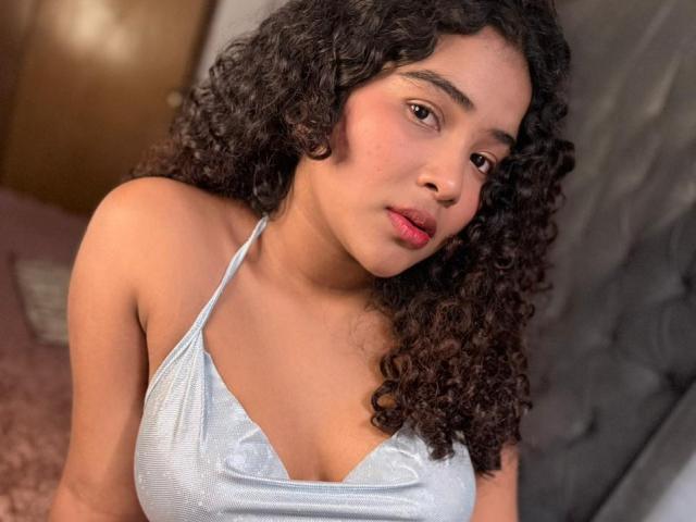 AnnyRestrepo with curly black hair, light brown skin, and red lipstick, wearing a shiny blue halter top, showing her small, cute tits, and a neutral, slightly pouty expression.