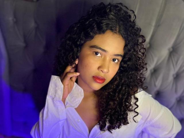 AnnyRestrepo with curly black hair, brown eyes, red lipstick, wearing a white button-up shirt, hand on cheek, looking at camera, gray tufted background, blue light on left.