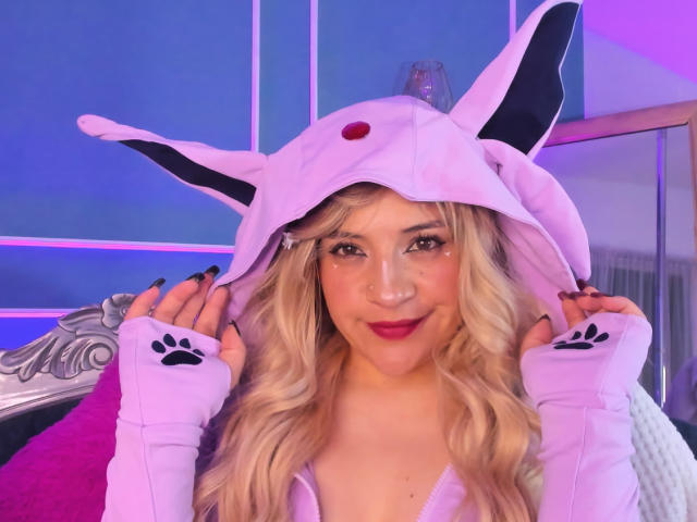 TaraTaylor with blonde hair, wearing a pink, cat-ear hooded cosplay outfit, purple paw gloves, and red lipstick, smiling with black nails, in a purple-lit room.