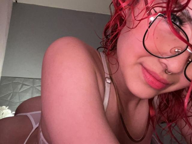 SharonFenix with curly red hair, glasses, nose ring, and pink lipstick, wearing white lingerie, showing her tits and smiling at the camera. She's leaning forward.