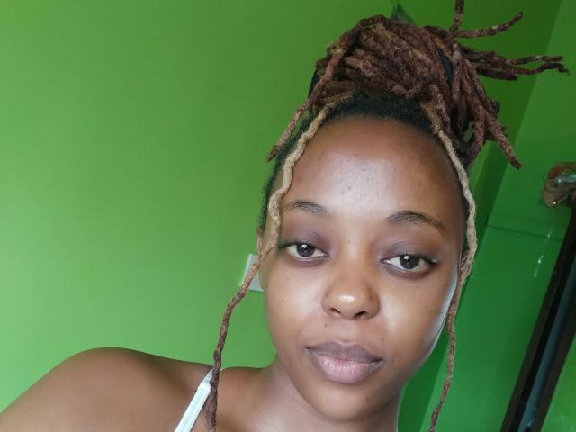 BlackElla has dark brown skin, small nose, and medium brown eyes with subtle makeup. She has long brown and blonde twist-up dreadlocks, and is wearing a white top.