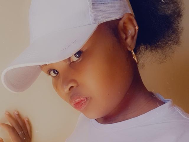 CorrieDee with a white cap, dark skin, and a small hoop earring in her left ear, wearing a white shirt, showing her pouty pink lips. Her hand is on a wall.