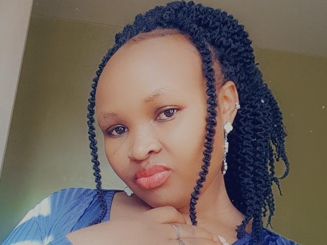 CorrieDee with dark curly braids, brown skin, pink lips, wearing a blue top, earrings, and a ring, looking at the camera with a slight pout.