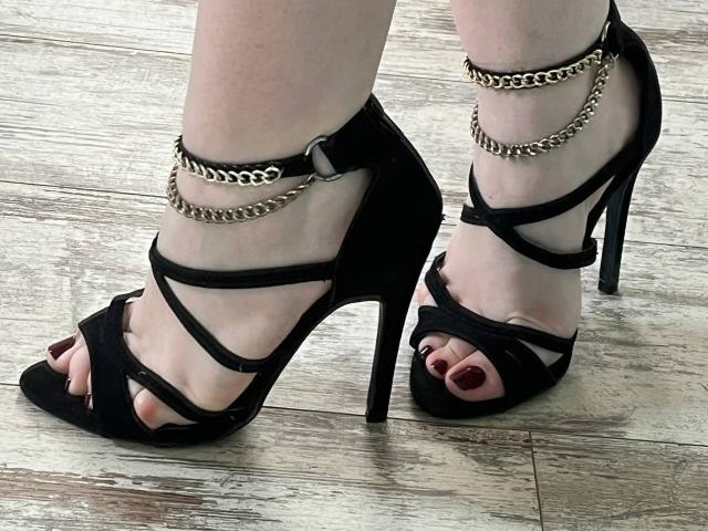 Valissia's feet in black high heels with strappy designs and gold chain ankle cuffs, showing red-painted toenails, standing on wooden floor. Her heels are super tall and sexy.