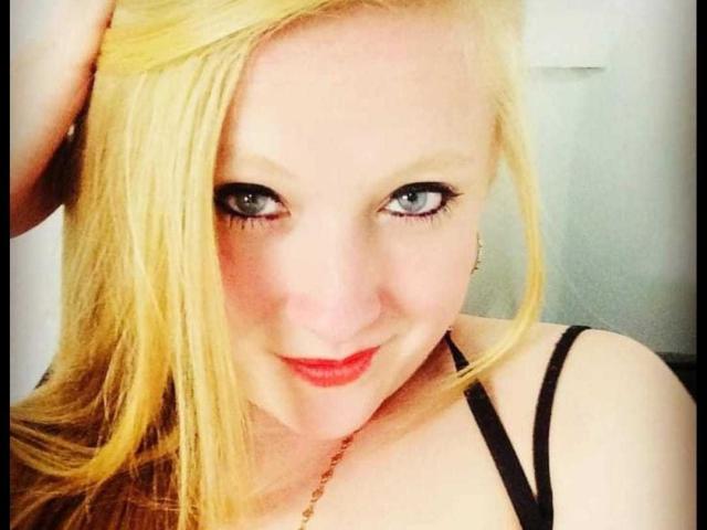 AliceCathyHot with blonde hair, blue eyes, red lipstick, and black strap dress, smiling at the camera, hand in hair, showing off her tits and cute face.