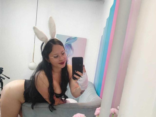 MaraVixen with black hair, glasses, white bunny ears, black lingerie, and gloves, taking a mirror selfie, showing her tits and ass, on a bed with pink and blue decor.