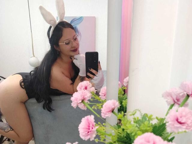 MaraVixen with long black hair, bunny ears, and glasses, showing her ass in a black thong, leaning on a gray chair, holding a phone, with pink flowers in foreground.