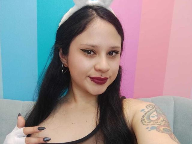 MaraVixen with black hair, wearing white bunny ears, red lipstick, and black eyeliner, showing off her tattooed arm and holding her tits. Her nails are painted black.