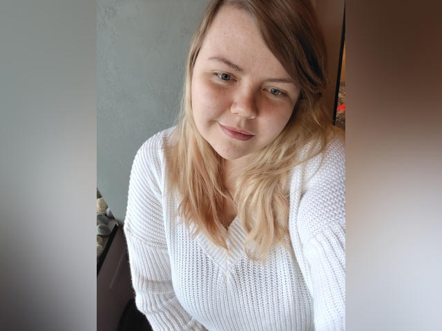 AsmiTKeyn with blonde hair, white waffle knit sweater, slightly smiling, standing between two white walls, looking at the camera, showing a cute, soft-ass face.