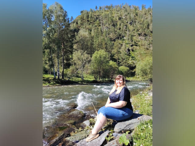 AsmiTKeyn with large tits, sitting on rocks by a flowing river, wearing a black shirt and blue jeans, smiling at the camera, surrounded by green trees and a forested hill.