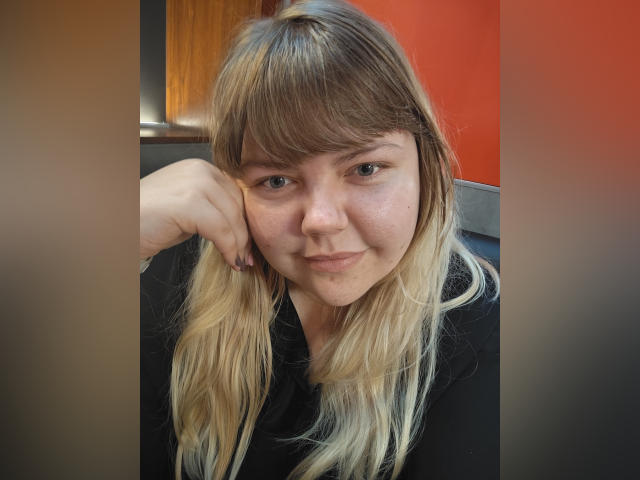 AsmiTKeyn has dirty blonde, shoulder-length hair, bangs, and a cute face with a slight smile. She's wearing a black top, resting her chin on her hand.