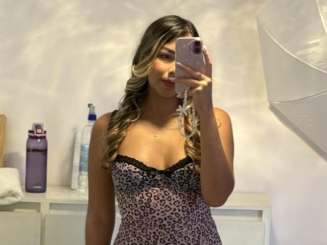 MaraCortez with long, wavy blonde hair, wearing a leopard print dress with black lace trim, holding a pink phone, taking a mirror selfie, showing her tits.