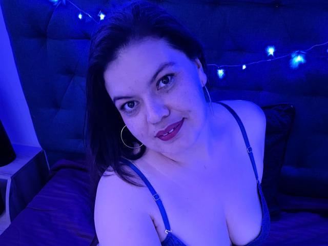 Gaiax69, eine kurvige Frau mit blauer BH, großen Brüsten, dunklem Haar, Reifenohrringen und rotem Lippenstift, lächelt in einem blau beleuchteten Schlafzimmer..