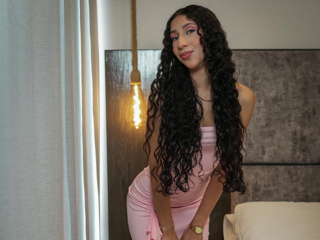 BeautifulShalom with long curly black hair, pink dress, and gold watch, leaning forward, showing her ass, with a sexy smile, and a hanging light bulb in the background.