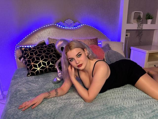 FelicityOnline, blonde, laying on bed, black dress, cleavage, red lipstick, tattoo on arm, blue LED strip, green quilt, black and gold pillow, white nightstand, potted plant.