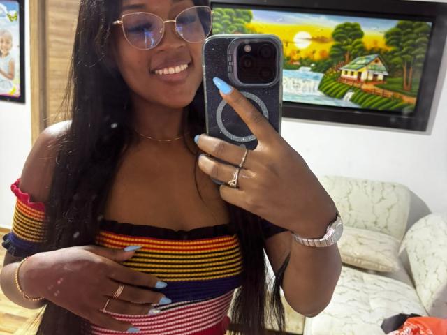 TaniaDoux with dark skin, long black hair, smiling, wearing glasses, off-shoulder striped top, blue nails, ring, and watch, taking a mirror selfie with a smartphone.