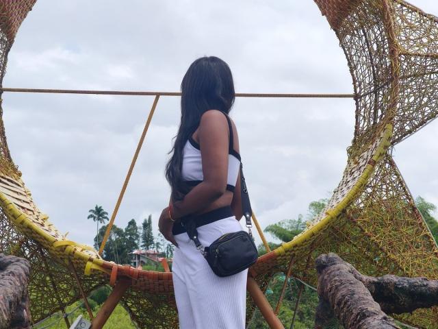 TaniaDoux with long black hair, wearing a white dress and black crossbody bag, standing in front of a yellow bamboo swing, looking away, back to the camera.