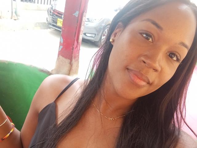 TaniaDoux has brown skin, long black hair, small gold earrings, and a subtle smile. She's wearing a black spaghetti strap top, a gold necklace, and colorful bracelets.