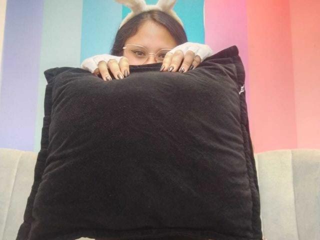 MaraVixen with glasses, black nails, holding a huge black pillow, wearing a white and gray long-sleeve shirt, and a bunny hat, sitting on a gray couch.