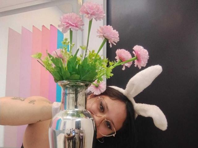 MaraVixen with glasses, pink carnations in a silver vase, white bunny ears, and a tattoo on her arm, peering over the vase, black background.