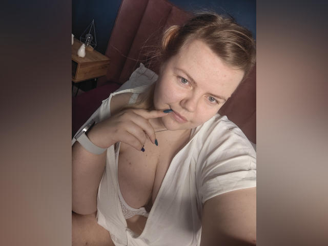 AsmiTKeyn with light skin, brown hair, and blue eyes, wearing a white shirt open to reveal her large, lace-covered tits. She's biting her finger with dark blue nails.