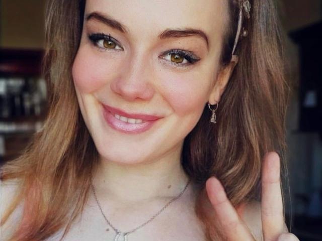 Noukonekox-hot has light brown hair, green eyes, and a cute smile. She's making a peace sign, showing off her small tits and pink lips. She wears a delicate necklace.
