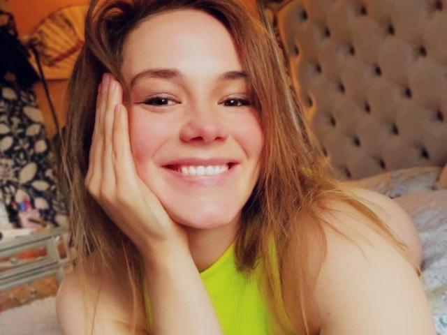 Noukonekox-hot with long brown hair, smiling, hand on cheek, wearing a neon green top, sitting on a bed, tufted headboard in background, soft lighting, cute, happy.