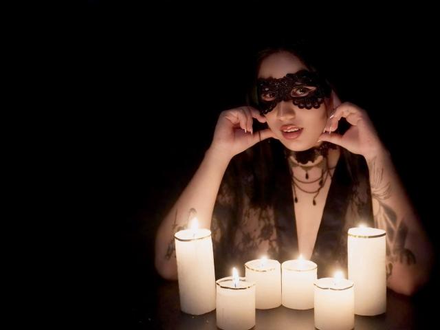 LiliethWolf with black mask, dark sheer robe, candles in front, hands near mask, light from candles on face, tattoo on right arm, dark background, serious expression.
